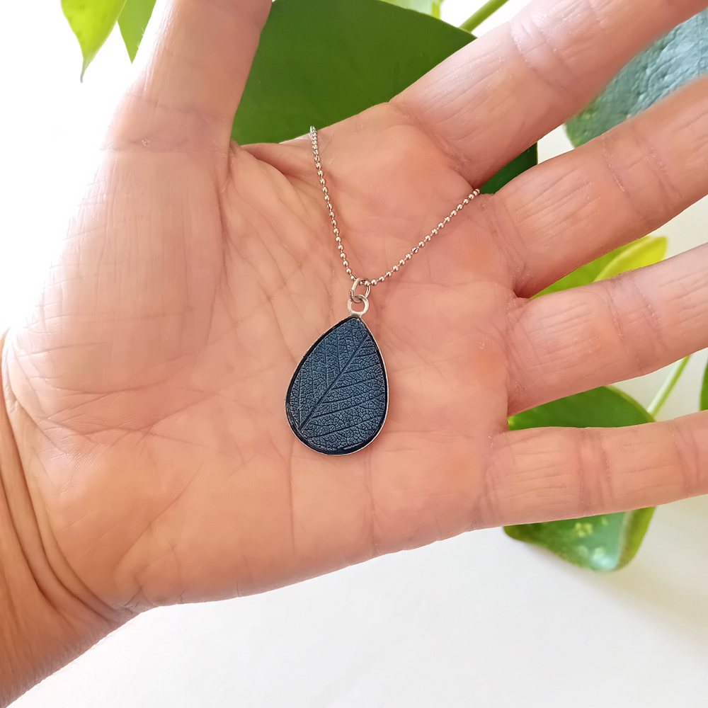 collier a planter par modibulle, sur chaine en acier inox, cabochon en forme de goutte de goutte de couleur bleu de prusse, contenant des graines d'ancolies des jardins, présenté dans la paume de la main