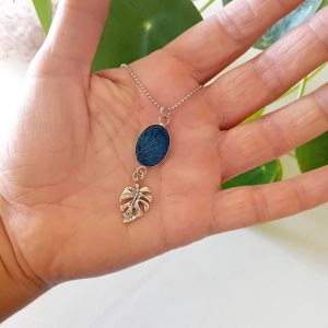 collier a planter, avec sa petite breloque en forme de feuille de monstera. Le médaillon est oval et de couleur bleu de Prusse et contient des graines d'ancolies des jardins. Par modibulle