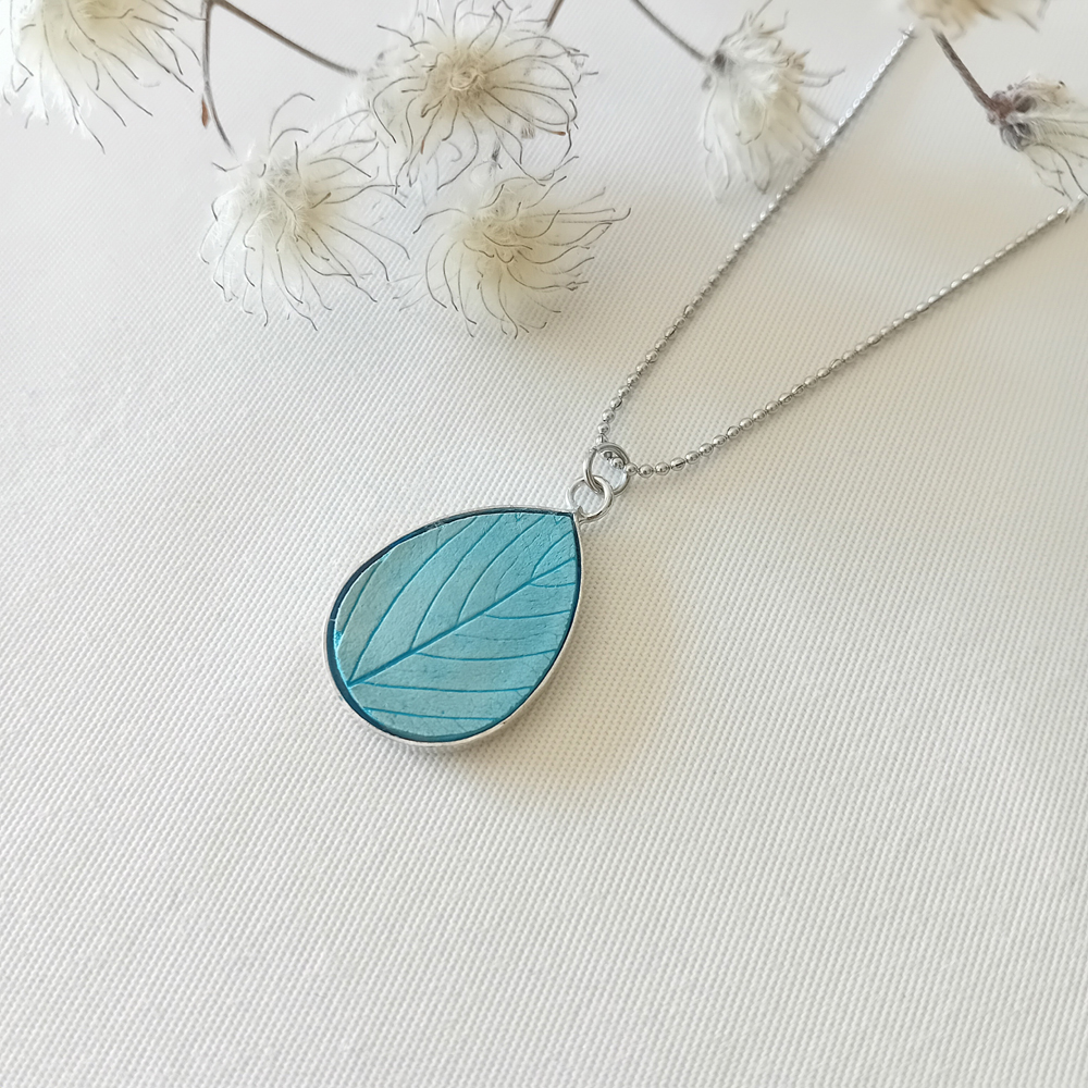 un collier à plater par modibulle, en forme de goutte et de couleur bleu azur, contenant des graines de fleur de nigelles
