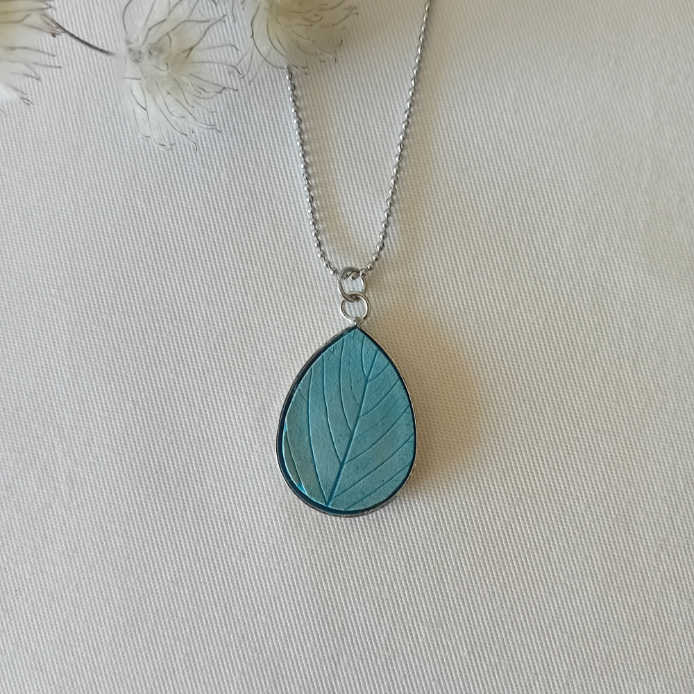 un collier à plater par modibulle, en forme de goutte et de couleur bleu azur, contenant des graines de fleur de nigelles
