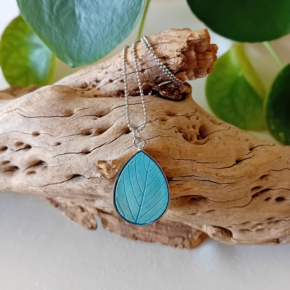 un collier à plater par modibulle, en forme de goutte et de couleur bleu azur, contenant des graines de fleur de nigelles