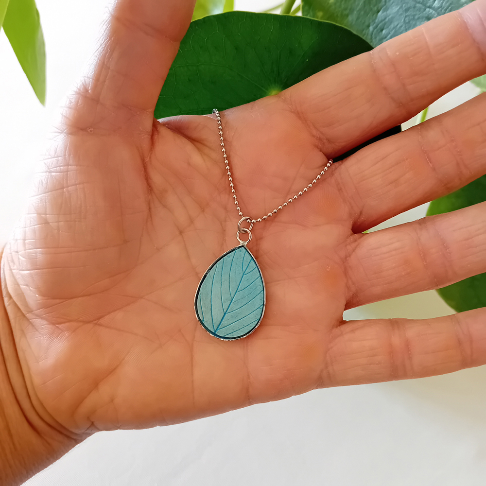 un collier à plater par modibulle, en forme de goutte et de couleur bleu azur, contenant des graines de fleur de nigelles